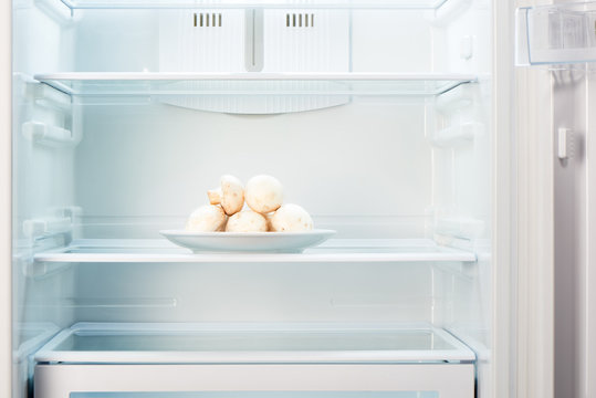 Fresh champignon mushrooms on white plate in open empty refrigerator