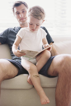 Man sitting on a sofa with his son, holding a book, reading.