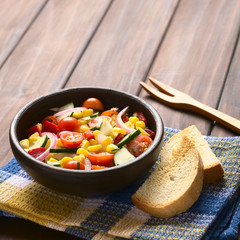 Vegetable salad made of sweet corn, cherry tomato, cucumber, red onion, red pepper, chives, photographed on dark wood with natural light (Selective Focus, Focus one third into the salad)