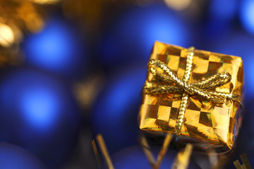 Close-up of a miniature gold gift box against a background of blue Christmas balls. Focus is on gift box.