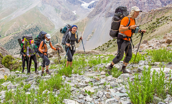 People Traveling In Mountains.
Large Group Of Tourists Of Different Sex Ethnic Nation Race Age Young And Old Man Woman Walking Up On Rocky Path With Green Grass Forest And Mountain Peaks Around