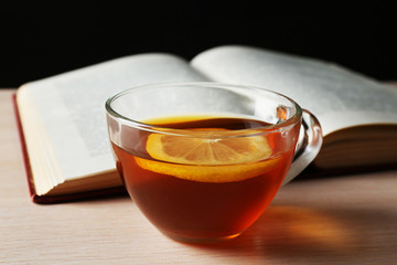 Glass cup of tea with book on wooden table on black background