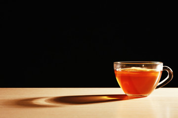 Glass cup of tea with piece of lemon on wooden table on black background