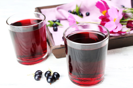 Glasses of fresh blackcurrant juice on wooden table near tray with flowers, closeup