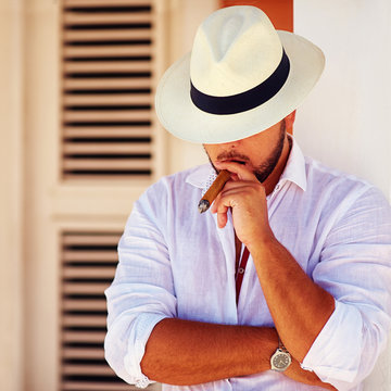 confident handsome man smoking cigar, while leaning on the wall