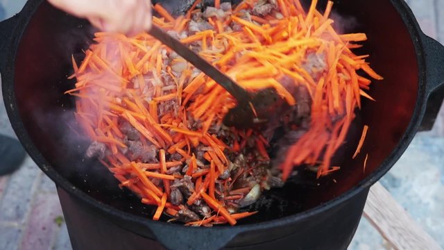 Uzbek national dish pilaf in a large cast-iron cauldron on the fire, add sliced red carrots, rice, lamb, mutton fat, the fat tail, sesame oil, onion, garlic, red hot chili pepper