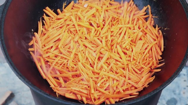Uzbek national dish pilaf in a large cast-iron cauldron on the fire, add sliced red carrots, rice, lamb, mutton fat, the fat tail, sesame oil, onion, garlic, red hot chili pepper