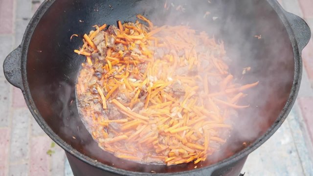 Uzbek national dish pilaf in a large cast-iron cauldron on the fire, add sliced red carrots, rice, lamb, mutton fat, the fat tail, sesame oil, onion, garlic, red hot chili pepper