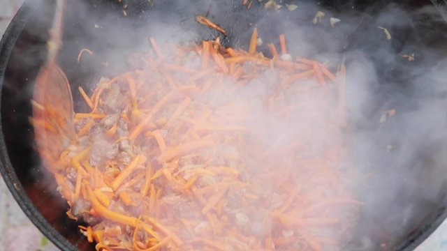 Uzbek national dish pilaf in a large cast-iron cauldron on the fire, add sliced red carrots, rice, lamb, mutton fat, the fat tail, sesame oil, onion, garlic, red hot chili pepper