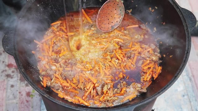Uzbek national dish pilaf in a large cast-iron cauldron on the fire, add sliced red carrots, pour cold water, rice, lamb, mutton fat, the fat tail, sesame oil, onion, garlic, red hot chili pepper