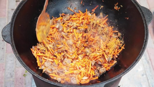 Uzbek national dish pilaf in a large cast-iron cauldron on the fire, sprinkled salt, cumin seeds, red carrots, rice, lamb, mutton fat, the fat tail, sesame oil, onion, garlic, red hot chili pepper