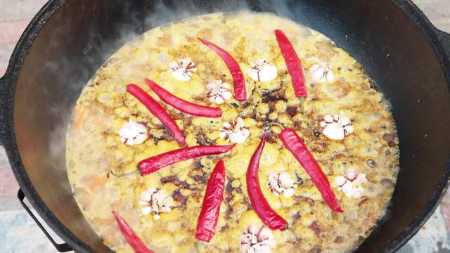 Uzbek national dish pilaf in a large cast-iron cauldron on the fire, sliced red carrots, cold water, add whole of garlic and red chili peppers, begins to boil, rice, lamb, mutton fat, the fat tail