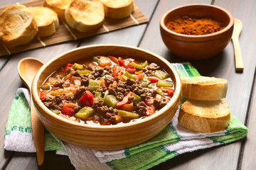 Vegan goulash made of soy meat (textured vegetable protein), capsicum, tomato and onion, served in wooden bowl accompanied by toasted bread, photographed with natural light