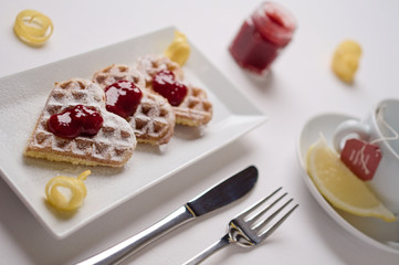 Heart shaped waffles, berry jam, powdered sugar served on rectangular plate with lemon slice, small jam jar and tea cup in background. Selective focus