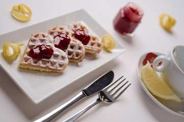 Heart shaped waffles, berry jam , powdered sugar served on  rectangular plate with tea cup, lemon slice, and small jar of berry jam in background