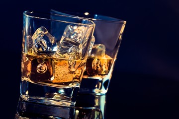 two glasses of whiskey with ice cubes on black background with light tint blue