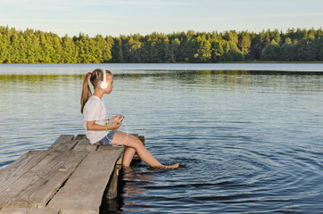 Fototapeta na wymiar ГАРМОНИЯ/ Девочка слушает музыку сидя на деревянном мостике на озере. 