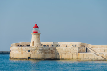 The lighthouse with red top of tower. The base of the lighthouse of yellow stone with embrasures. The lighthouse is located on background of the  blue sky and sea. © annaleish