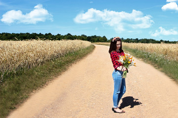 Portrait of young beautiful girl in the red shirt istanding on the road . Happy romantic girl in the wheat field. Pretty young girl with summer bouquet of flowers. Free happy woman