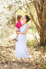 pregnant mother in white dress holding kissing her daughter in pink clothes on spring summer day in park outside among blooming trees 