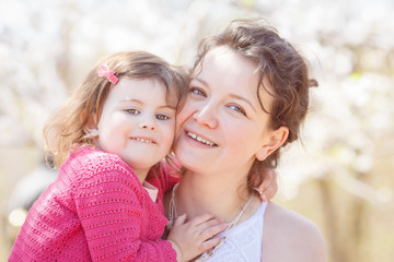 Obraz premium Close up portrait of caucasian mother in white dress holding hugging her daughter in pink clothes kissing her in cheek on sunny spring summer day in the park outside 