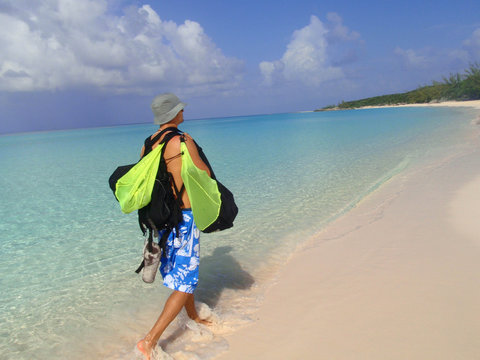 Empty sandy beach and light blue water. A man walking towards the bay  early in the morning carrying snorkeling equipment. Little San Salvador Island, also known as Half Moon Cay, Bahamas.