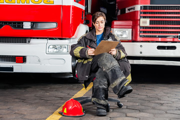 Fototapeta premium Firewoman Writing On Clipboard While Sitting Against Trucks