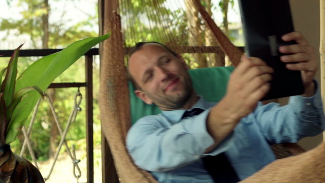 Young businessman chatting on tablet computer while lying on hammock 
