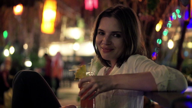 Young, happy woman raising toast to camera and drinking cocktail in cafe at night
