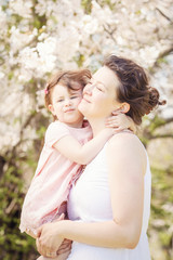 Fototapeta premium Portrait of caucasian pregnant mother in long white dress holding and kissing her daughter in pink clothes on spring summer day in park outside among blooming cherry trees 
