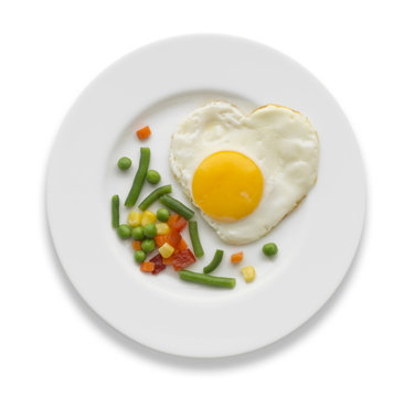 Fried egg in a shape of the heart and some vegetables on a white plate.