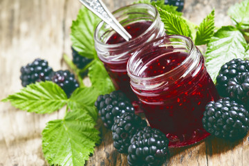 Two jars of blackberry jam, fresh berries and green leaves in an