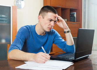 Sad guy staring at financial documents in laptop at table in hom