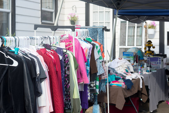 A rack of used clothing at a neighborhood yard sale with a variety of items in the background.