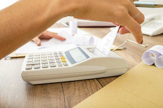 Bookkeeper doing calculations on an adding machine checking the totals against those in the journals, close up of her hands and the machine on a wooden desk.