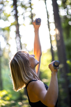 Young blond woman working out outdoors in woodland lifting weights extending the dumbbells to the glow of the sun through the trees with a happy smile.