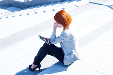 Redhead attractive woman sitting on the street on the stairs with tablet