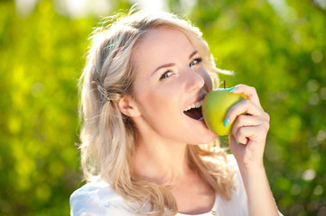 Smiling blonde girl biting green apple outdoors. Looking at camera. Healthy eating. 