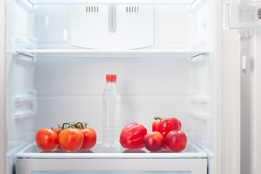 Branch of red tomatoes, two red peppers, two two-colored orange and red peaches and a bottle of water on shelf of open empty refrigerator. Weight loss diet concept.