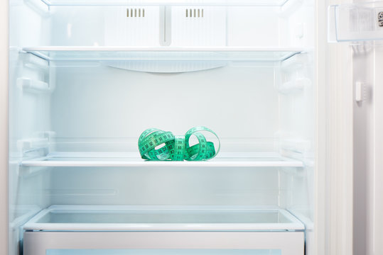 Green measuring tape on shelf of open empty refrigerator. Weight loss diet concept.