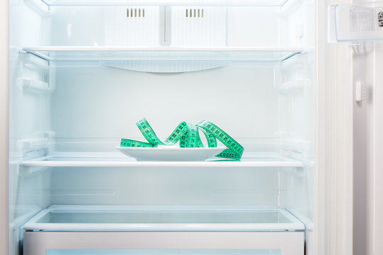 Green measuring tape on white plate in open empty refrigerator. Weight loss diet concept.
