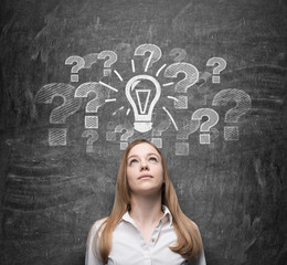 Beautiful young woman is thinking about undiscovered issues. Question marks and a light bulb are drawn on the black chalkboard behind the person.