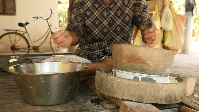 Woman sat cross-legged using hand-turned millstone to grind wet rice for making soaked rice flour