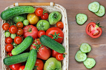 Свежие огурцы, красные и зеленые помидоры  на столе лежат в корзинке, вид сверху. Помидоры зрелые и недозрелые. 