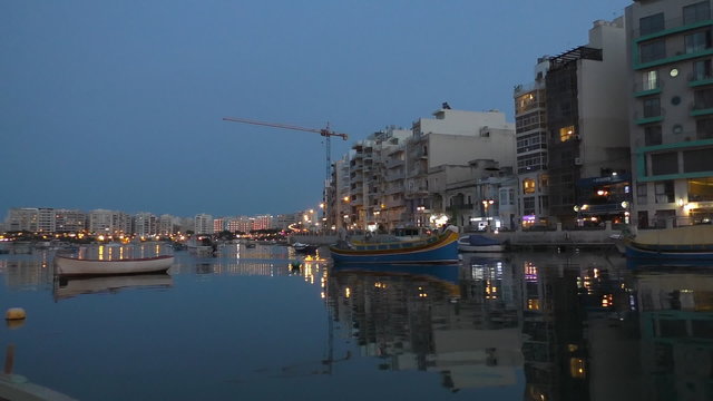 Beautiful Malta island harbor city night scene.Traditional Maltese boat coming toward camera at Spinola bay St Julians city in the evening