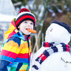 Funny kid boy in colorful clothes making a snowman, outdoors