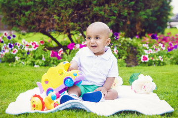 Portrait of cute adorable little indian South Asian infant boy in white shirt sitting on ground with toys in park green grass blanket outside on bright summer day smiling and playing