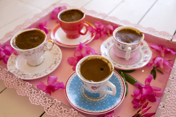 Four small cups of traditional foamy Turkish Coffee serving on a colorful flowery pink tray on top of a white wooden table in low angle view