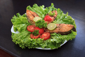A piece of fried fish fillett served on blue glass table with lettuce, lime, tomato and herbs
