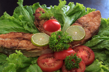 A piece of fried fish fillett served on blue glass table with lettuce, lime, tomato and herbs